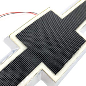 Puede incluir: Un emblema de Chevrolet en forma de pajarita con un panel central negro texturizado con patrón de rejilla, rodeado por un borde blanco iluminado y un marco exterior de cromo pulido, con cables eléctricos finos rojos, negros y blancos.