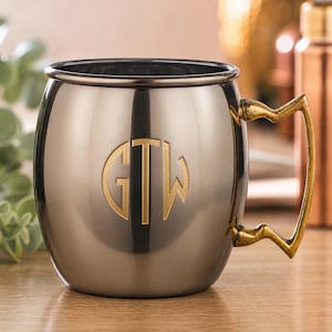 Pode incluir: Uma caneca Moscow Mule em aço inoxidável com uma alça dourada e um monograma dourado com as letras GTW. A caneca tem um acabamento metálico brilhante e assenta numa superfície de madeira. A caneca foi concebida para bebidas frias.