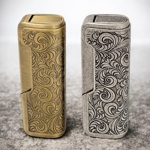 May include: Two decorative lighters are shown, one gold and one silver. Both lighters have an elaborate, swirling floral design. The gold lighter has a brushed finish, while the silver lighter has a matte finish. The lighters are side-by-side.