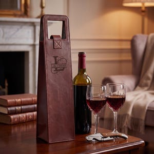 May include: A brown leather wine bottle carrier with a handle, personalized with the name "LACOMBE". A wine bottle, two wine glasses, and a corkscrew are on a wooden table. The glasses are filled with red wine.