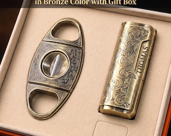 May include: A bronze-colored cigar lighter and V-cutter gift set in a presentation box. The lighter features an engraved scrollwork pattern and the name THOMAS. The V-cutter has a matching ornate design. Text reads: Personalized Cigar Lighter & V-Cutter Gift Set in Bronze Color with Gift Box.