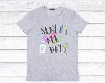 Może przedstawiać: Szara koszulka z nadrukiem "SLAY ALL DAY" w kolorze czarnym. Tekst jest otoczony kolorowymi ilustracjami pędzli, kredek, nożyczek, kleju i odcisków dłoni.