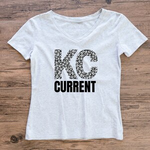 May include: A gray v-neck t-shirt with the letters "KC" made up of soccer balls and the word "CURRENT" printed in black below.
