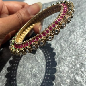 Peut inclure: Un bracelet jonc doré orné d'une rangée de pierres magenta ovales alternant avec des pierres claires circulaires serties dans un métal travaillé. La surface intérieure du jonc présente un motif géométrique texturé et répétitif.