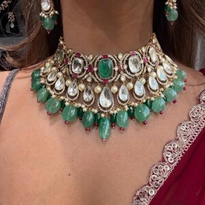 May include: A gold-toned necklace and matching earrings set with emerald green and clear gemstones, and small red accents. The necklace features large emeralds and teardrop-shaped clear stones.