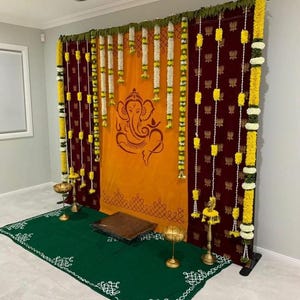 May include: A decorative backdrop with maroon and orange fabrics, adorned with floral garlands. The orange fabric features a Ganesha illustration. A green rug with white patterned edges and brass lamps are included.