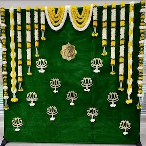 May include: A green backdrop adorned with garlands of yellow and white flowers. Gold lotus flower cutouts and a gold emblem are attached to the backdrop. The garlands are arranged vertically and horizontally, creating a festive display.