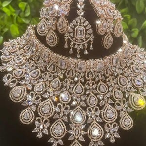 Puede incluir: Un conjunto de joyas de novia en tono oro rosa que incluye collar, pendientes y tocado. El conjunto está incrustado con cristales transparentes y brillantes de varias formas, incluyendo lágrimas y cuadrados. Las joyas se exhiben sobre un fondo oscuro.
