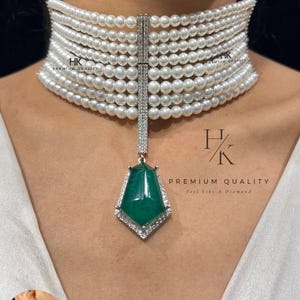 Może przedstawiać: Perłowy naszyjnik typu choker z diamentowym zapięciem i zieloną zawieszką z kamienia szlachetnego. Naszyjnik wykonany jest z wielu sznurów białych pereł. Zawieszka to zielony kamień szlachetny z diamentową obwódką. Widoczny jest tekst "HK PREMIUM QUALITY".