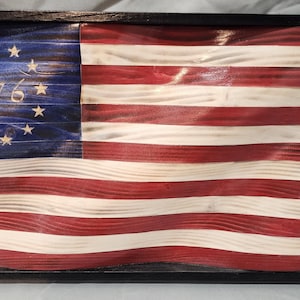 May include: A wooden American flag with a wavy design, framed in black. The flag features red and white stripes and a blue field with white stars and the year "1776".