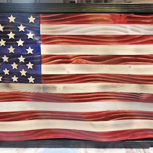 Op de afbeelding: Een houten Amerikaanse vlag met een distressed afwerking. De vlag heeft rode en witte strepen en een blauw veld met witte sterren. De vlag is omlijst in een zwart houten frame.