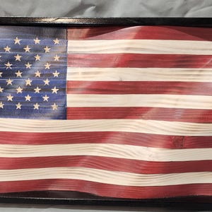 Könnte beinhalten: Eine gerahmte amerikanische Flagge aus Holz mit wellenförmigem Design. Die Flagge zeigt rote und weiße Streifen und ein blaues Feld mit weißen Sternen. Die Holzmaserung ist sichtbar. Der Rahmen ist schwarz.