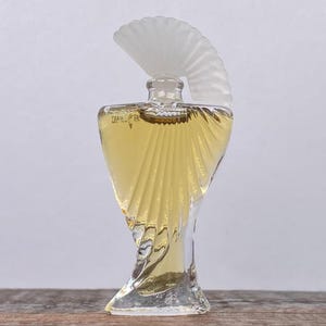 May include: A clear glass perfume bottle with a fan-shaped stopper. The bottle has a unique, sculpted design with a yellow liquid inside. The bottle is on a wooden surface against a white background.