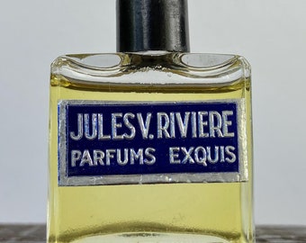 May include: A vintage perfume bottle of Jules V. Riviere Parfums Exquis. The clear glass bottle has a dark cap and a blue and silver label. The liquid inside is yellow. The bottle is rectangular and sits on a wooden surface.