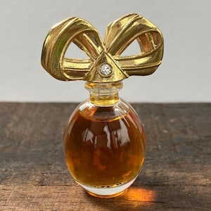 Può includere: Piccola bottiglia di profumo rotonda riempita di liquido ambrato. Il tappo è dorato e a forma di fiocco, ornato da un brillante trasparente. La bottiglia è appoggiata su una superficie di legno.