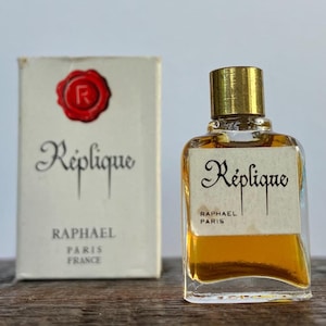May include: A small glass bottle of Replique perfume by Raphael Paris. The bottle has a gold cap and a label with the brand name and city. The bottle is sitting on a wooden surface.