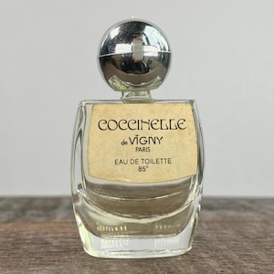 May include: A small glass bottle of perfume with a silver cap. The label reads "Coccinelle de Vigny Paris Eau de Toilette 85°".