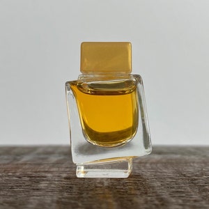May include: A small, clear glass bottle with a gold top, filled with a yellow liquid. The bottle has a unique, angular design.