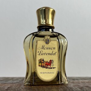 May include: A clear glass bottle of Mousson Lavende cologne with a gold cap and label. The label features a gold crest and a picture of a horse-drawn carriage. The text on the label reads "Mousson Lavende" and "Mit der Postkutsche".