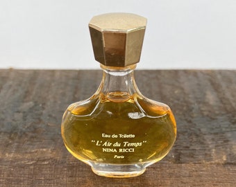 May include: A small, clear glass bottle of perfume with a gold cap. The bottle is shaped like a teardrop and has the text "Eau de Toilette" "L'Air du Temps" "Nina Ricci" "Paris" on the label.