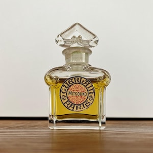 Może przedstawiać: Przezroczysty szklany flakon perfum z ozdobnym korkiem. Butelka zawiera płyn w kolorze złotym i posiada etykietę z napisem "GUERLAIN PARIS MITSOUKO". Butelka ma 7,6 cm wysokości.