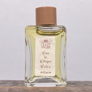 May include: A rectangular glass bottle of Eau de Cologne Extra by Adam, with a brown cap. The label features text and a crest design. The bottle is filled with a light yellow liquid.
