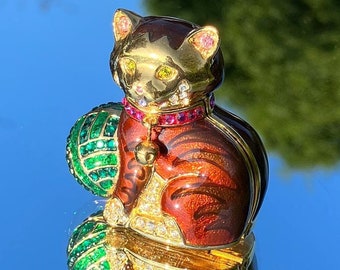 以下が含まれることがあります： 緑色のボールを持った、金、赤、緑の宝石で飾られた猫の置物。猫はピンクの首輪に鈴をつけています。