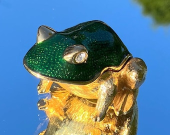 May include: A small, green and gold frog trinket box with a single rhinestone eye. The frog is sitting on a reflective surface.