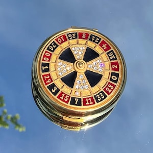 Peut inclure: Un miroir compact doré avec un design de roue de roulette noir et blanc. La roue comporte des numéros rouges et noirs et des diamants blancs autour du bord.