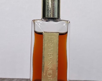 Può includere: Un flacone di profumo rettangolare in vetro con un tappo color oro e un'etichetta con la scritta "Cinnabar". Il flacone contiene un liquido ambrato scuro. Il flacone è su una superficie di legno.