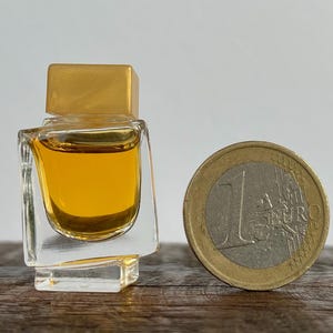 May include: A small, clear glass bottle with a gold top filled with a yellow liquid. The bottle is sitting on a wooden surface next to a euro coin.