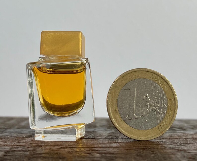 May include: A small, clear glass bottle with a gold top filled with a yellow liquid. The bottle is sitting on a wooden surface next to a euro coin.
