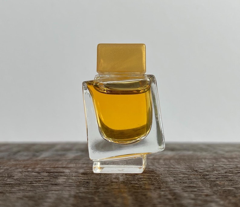 May include: A small, clear glass bottle with a gold top, filled with a yellow liquid. The bottle has a unique, angular design.