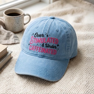 May include: Light blue baseball cap with the embroidered text "Over Stimulated & Under Caffeinated" in pink. The cap is sitting on a cream-colored surface, with a coffee mug and books in the background.