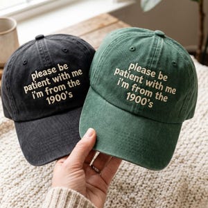 May include: Two baseball caps, one in charcoal black and one in forest green, both featuring embroidered cream-coloured text that reads: please be patient with me i'm from the 1900's. Each cap has a structured crown, a curved brim, and eyelets.