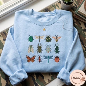 Peut inclure: Un sweat-shirt bleu clair à col rond orné d'une grille brodée de douze insectes colorés, dont un papillon monarque, une coccinelle, une abeille et divers coléoptères. Un collier argenté délicat avec un petit pendentif rond repose sur l'encolure.