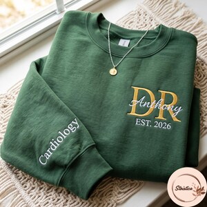 May include: A forest green crewneck sweatshirt featuring gold embroidered initials DR with the name Anthony in white script and EST. 2026 below. The sleeve is embroidered with the word Cardiology in white. A gold pendant necklace rests on the neckline.
