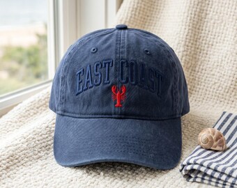 May include: A navy blue baseball cap with the embroidered text "EAST COAST" in a matching color. A red lobster is embroidered below the text. The cap is sitting on a cream-colored blanket next to a folded blue and white striped cloth.