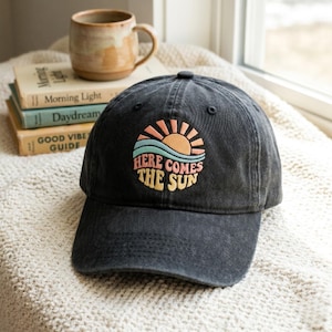 Puede incluir: Gorra de béisbol negra con visera curva. La gorra presenta un diseño bordado de un sol, olas y el texto "HERE COMES THE SUN" en una fuente retro. Una taza de cerámica y libros están en el fondo.