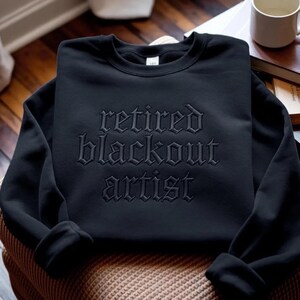 May include: A black long-sleeve crewneck sweatshirt featuring the text "retired blackout artist" embroidered in a matching black thread across the chest in a gothic-style font. The garment has a ribbed collar, cuffs, and hemline.