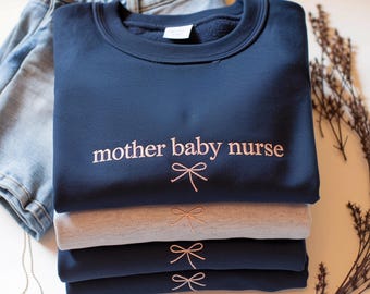 May include: A stack of navy blue and gray sweatshirts with the embroidered text "mother baby nurse" and bow designs. The sweatshirts are folded neatly, with a pair of blue denim shorts in the background.