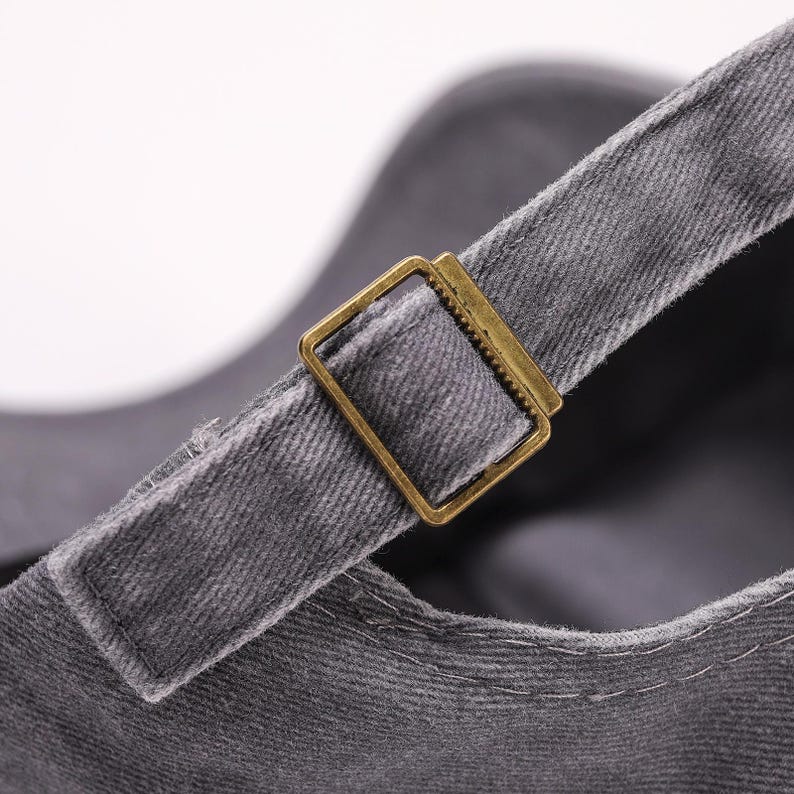 May include: Close-up of a gray baseball cap with a brass-colored metal buckle on the adjustable strap. The cap is made of a textured fabric, and the buckle is rectangular.