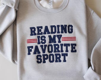 May include: A light gray sweatshirt with the embroidered text "READING IS MY FAVORITE SPORT" in navy blue block letters. Two red stripes are under the words "IS MY". The sleeves are folded up.