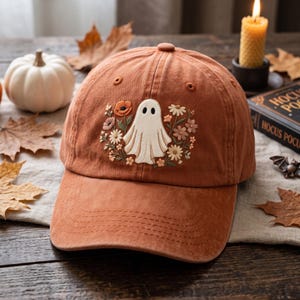 May include: A rust-colored baseball cap featuring an embroidered design of a white ghost surrounded by a variety of colorful flowers including orange poppies and small white daisies, with a button on top and stitched detailing on the curved brim.