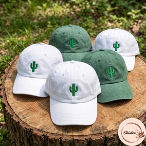 May include: A collection of five baseball caps in white and forest green, each featuring a small, embroidered green cactus icon centred on the front panel. The caps have a structured crown, a curved brim, and a button at the top centre.