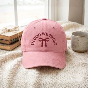 Peut inclure: Casquette de baseball rose poudré avec l'inscription "IN GOD WE TRUST" au-dessus d'un motif de nœud bordeaux. Le chapeau est en tissu doux et possède une visière incurvée. La casquette est présentée sur une couverture crème.