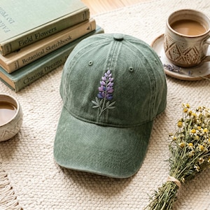 Könnte beinhalten: Eine salbeigrüne Baseballkappe mit einem lila und weißen gestickten Blumendesign. Die Kappe liegt auf einem cremefarbenen Teppich neben Büchern und einer Kaffeetasse. Ein kleiner Strauß Trockenblumen ist ebenfalls zu sehen.