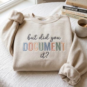 Könnte beinhalten: Ein beigefarbenes Sweatshirt mit Rundhalsausschnitt und gesticktem Text. Die Wörter but did you sind in dunkelgrauer Schreibschrift gehalten, während das Wort DOCUMENT in Blockbuchstaben in hellblau, pfirsich, salbeigrün und blau erscheint, gefolgt von it? in Grau.
