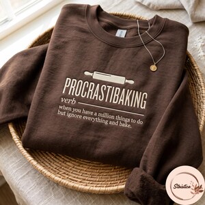 May include: A dark brown crewneck sweatshirt featuring a white graphic of a rolling pin and the text: PROCRASTIBAKING verb when you have a million things to do but ignore everything and bake. A thin gold chain necklace with a small round pendant rests on top.