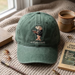 May include: A sage green baseball cap featuring an embroidered graphic of a fish wearing a tuxedo and reading a book. Below the fish, the text o-fish-iant is embroidered in white thread. The cap has a curved brim and a structured front panel.